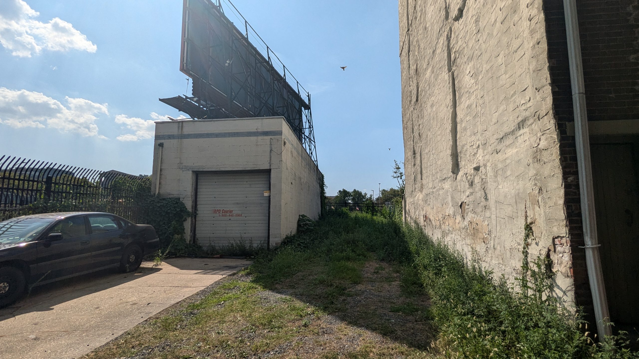 narrow vacant lot covered in grass and gravel squeezed between a single car garage with a car parked in the driveway on one side, and the bare side wall of a larger building on the other side. The backside of a billboard is visible behind the garage.